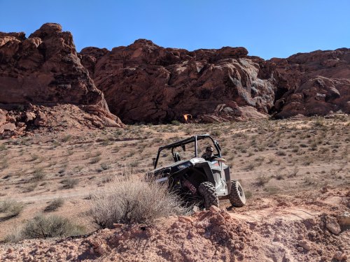 A side-by-side vehicle climbs a rocky terrain in a desert landscape with red rock formations in the background.
