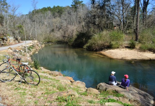 Two people sit on rocks by a calm river, with a bicycle nearby and trees lining the bank on a sunny day.
