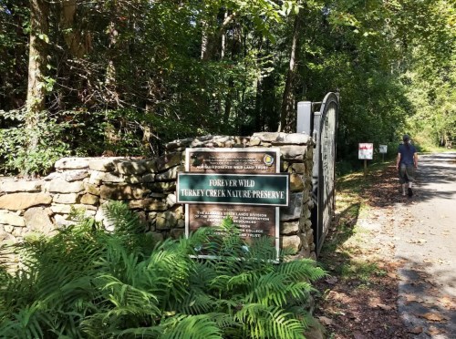 A stone entrance sign for Turkey Creek Nature Preserve, surrounded by trees and ferns, with a path leading into the woods.