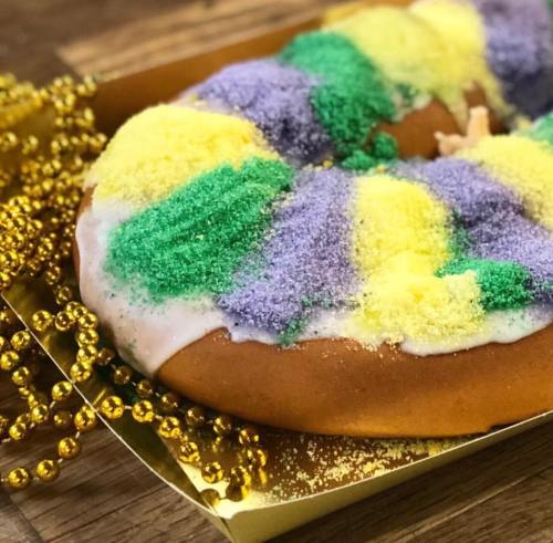 A colorful king cake topped with purple, green, and yellow sugar, surrounded by gold beads on a wooden surface.