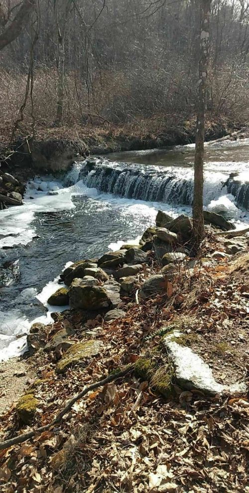 A serene river with a small waterfall, surrounded by rocky banks and bare trees in a winter landscape.
