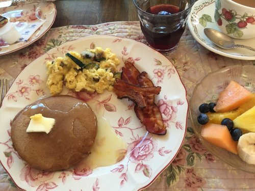 A plate with pancakes topped with butter, scrambled eggs, bacon, and a side of fresh fruit and a glass of juice.
