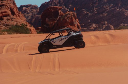 A side-by-side off-road vehicle climbs a sandy dune with rocky formations in the background.