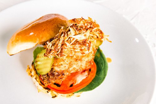 A close-up of a fried chicken sandwich with pickles, tomato, and slaw on a soft bun, served on a white plate.