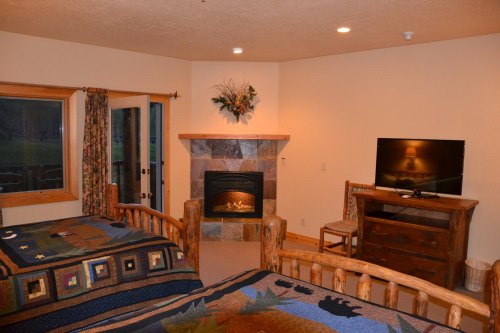 Cozy bedroom with two log beds, a stone fireplace, TV, and rustic decor, featuring warm lighting and natural elements.