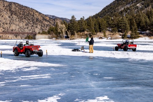 Two off-road vehicles on a frozen lake, with a person waving a flag and mountains in the background.
