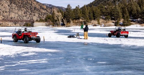 Two off-road vehicles on a frozen lake, with a person waving a flag and mountains in the background. Snowy landscape.