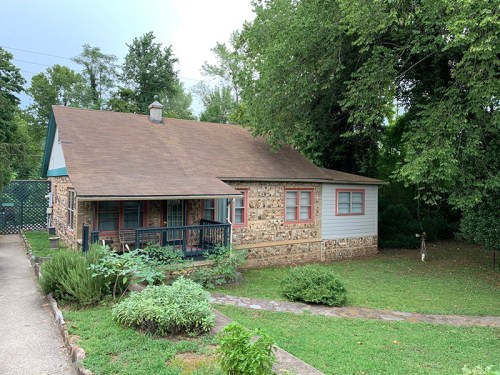 A cozy stone house with a brown roof, surrounded by greenery and trees, featuring a small porch and garden area.