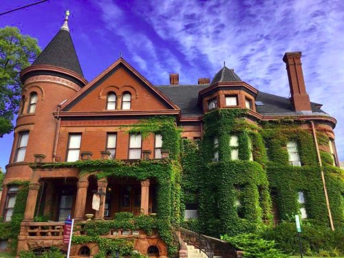 A large, ivy-covered brick mansion with turrets, set against a blue sky with wispy clouds.