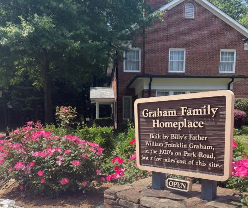 Sign for Graham Family Homeplace with a brick house and blooming flowers in the background, indicating it's open for visitors.