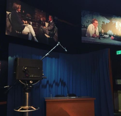 A television studio set with a camera, blue curtain, and screens showing interviews with two hosts.