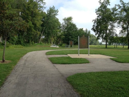 A winding path in a park leads to a historical marker, surrounded by trees and grassy areas.