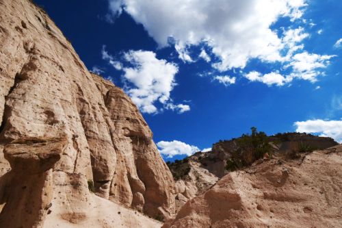 A rocky landscape with towering cliffs under a bright blue sky dotted with fluffy white clouds.