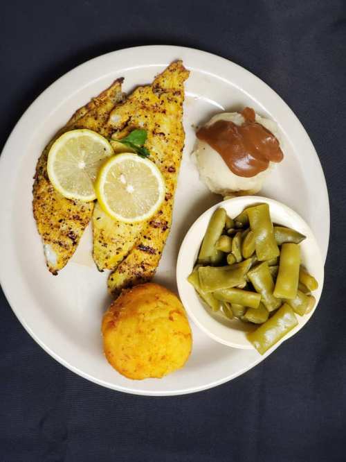 A plate with grilled fish fillets topped with lemon, cornbread, mashed potatoes with gravy, and green beans.