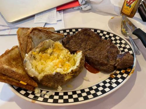 A plate with a grilled steak, a baked potato filled with cheese, and toasted bread, set on a checkered platter.