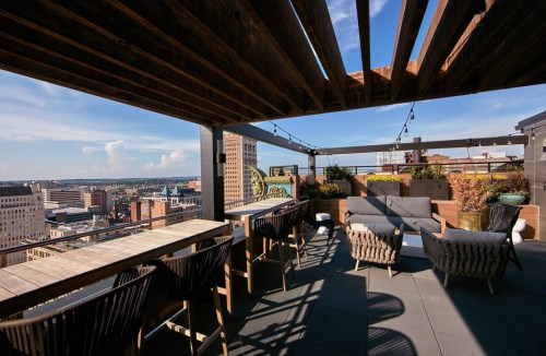 Rooftop terrace with seating and city skyline views, featuring wooden beams and potted plants under a clear blue sky.