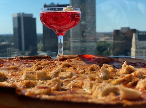 A close-up of a pizza with toppings, alongside a red cocktail, with a city skyline in the background.