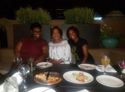 Three women smiling at a dining table with food and drinks, surrounded by greenery in an outdoor setting.