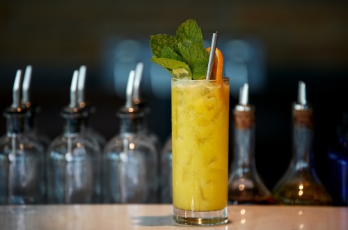 A tall glass of yellow cocktail garnished with mint and an orange slice, set against a blurred bar background.