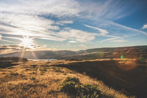 A scenic landscape featuring rolling hills, a river, and a bright sun setting against a blue sky with wispy clouds.
