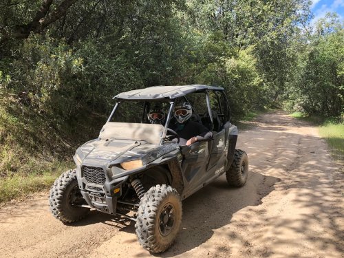 A side-by-side off-road vehicle on a dirt path, surrounded by trees, with two people wearing helmets inside.