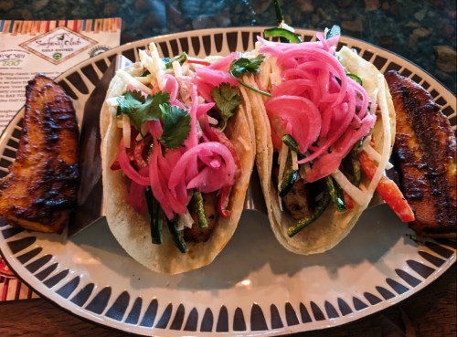 Two tacos filled with grilled meat, topped with pickled onions and cilantro, served with a side of grilled plantains.
