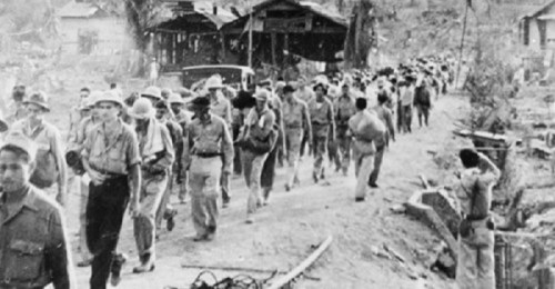 Black and white photo of a long line of soldiers marching along a dirt road, with buildings in the background.
