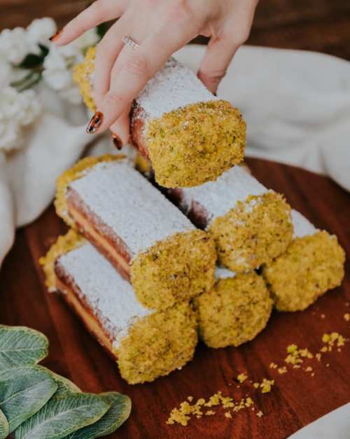 A hand reaches for a stack of pistachio-coated pastries dusted with powdered sugar on a wooden platter.