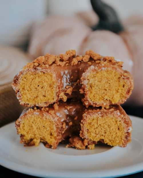 A stack of pumpkin donuts topped with crumbled cookies on a white plate, with decorative pumpkins in the background.