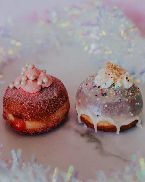 Two colorful donuts on a marble surface, one with pink sprinkles and pearls, the other with white icing and colorful sprinkles.