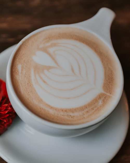 A close-up of a latte with a heart and leaf design on the frothy surface, served in a white cup on a saucer.