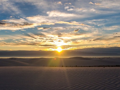 A stunning sunset over rolling sand dunes, with vibrant clouds and a golden sun illuminating the horizon.