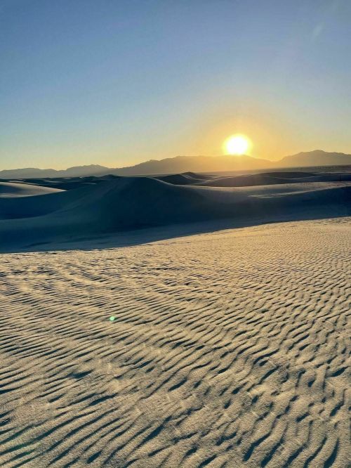 A serene desert landscape at sunset, with rippled sand dunes and mountains in the background.