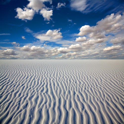 A vast white landscape with rippled sand under a bright blue sky filled with fluffy clouds.
