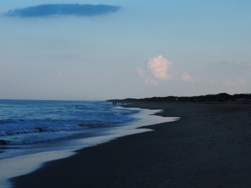 A serene beach at dusk, with gentle waves lapping the shore and a few distant figures walking along the water's edge.
