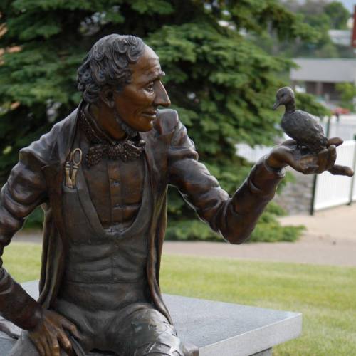 A bronze statue of a man in a suit holding a small bird on his hand, seated on a stone bench in a park.
