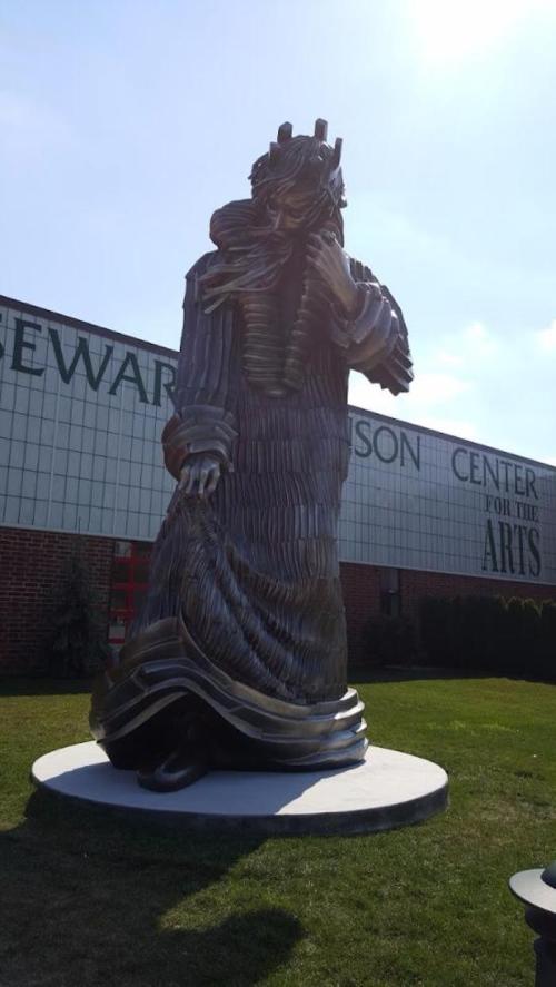 A large bronze sculpture of a figure in a flowing garment stands outside a building, with grass and blue sky in the background.