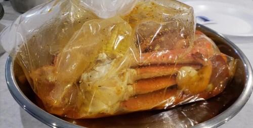 A sealed plastic bag filled with seasoned crab legs, resting in a metal bowl.