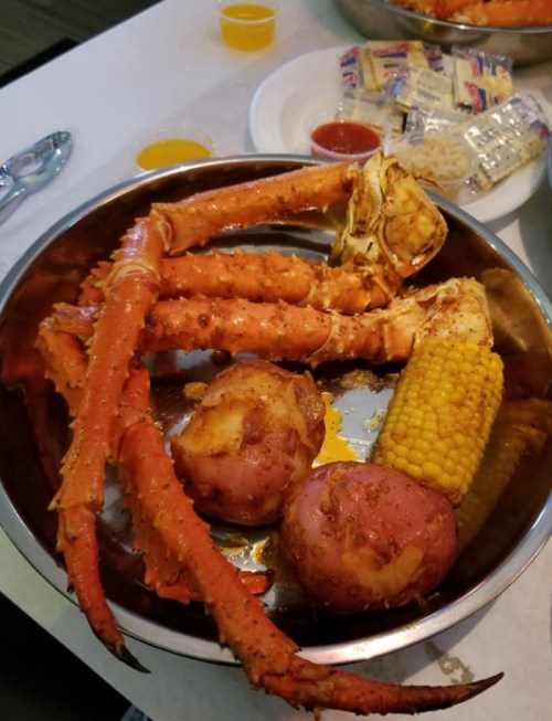 A metal bowl filled with crab legs, red potatoes, and corn, accompanied by dipping sauces and crackers on the side.