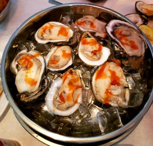 A metal bowl filled with fresh oysters on ice, topped with a drizzle of sauce.