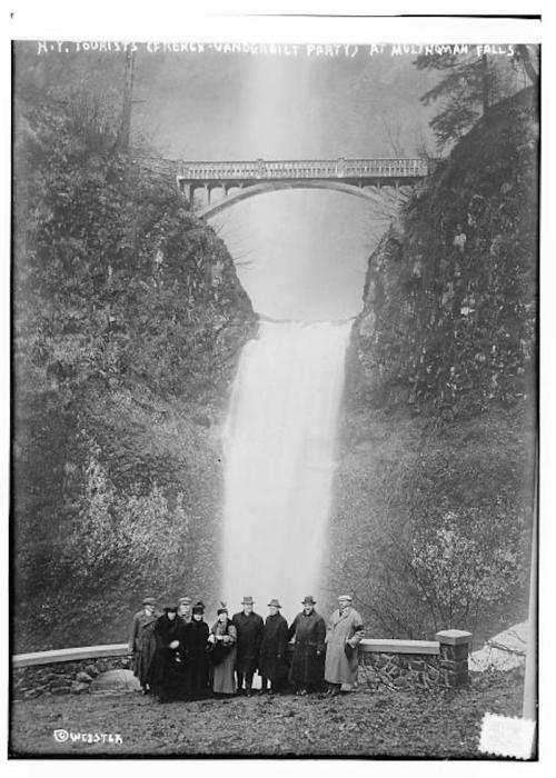 A group of tourists poses in front of Multnomah Falls, with a bridge and waterfall in the background.