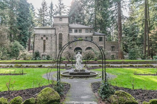 A stone building surrounded by trees, with a garden featuring a statue in the foreground.