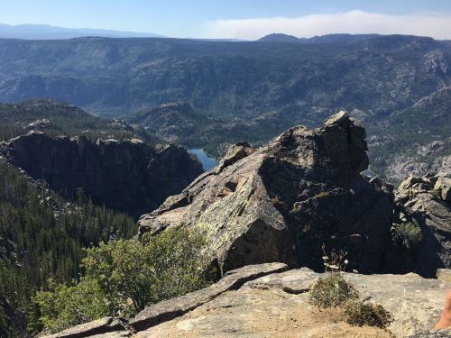 A panoramic view of rugged mountains and a serene lake surrounded by dense forests under a clear blue sky.