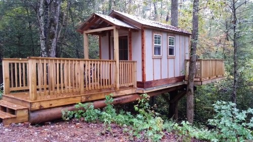 A small wooden treehouse with a porch, elevated among trees in a forested area.