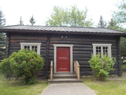 A rustic log cabin with a red door, surrounded by greenery and a pathway leading to the entrance.
