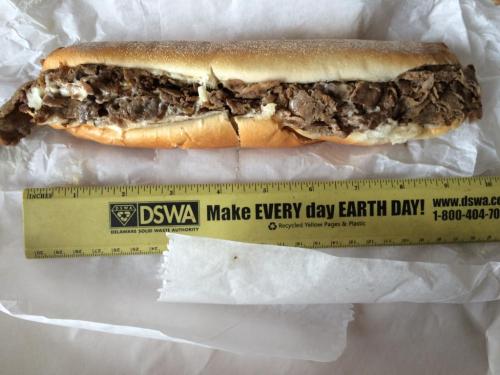 A long sandwich filled with sliced beef, placed on white paper next to a ruler for size reference.