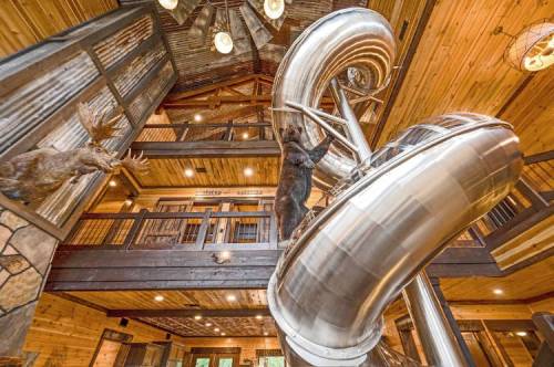 A large metal slide spirals down from a wooden loft in a rustic cabin, with a bear statue nearby.