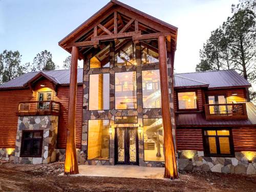 A modern log cabin with large glass windows, stone accents, and wooden beams, surrounded by trees at dusk.