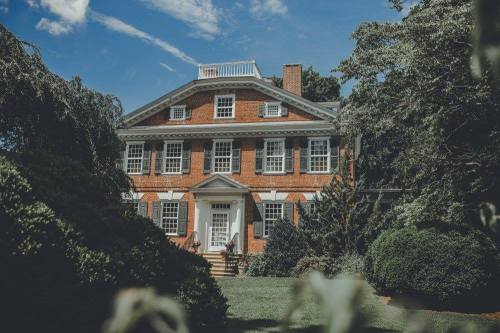 A large brick house with white trim, surrounded by lush greenery and under a clear blue sky.