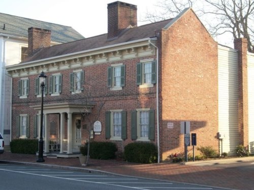 Historic brick house with green shutters, a porch, and a chimney, located on a street with parked cars.